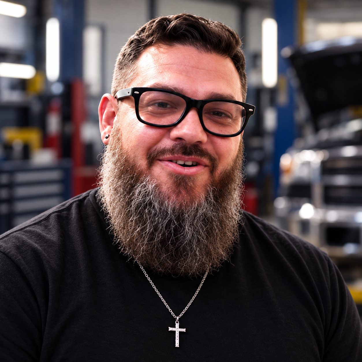 Bearded man in glasses, black shirt and cross necklace smiling inside a shop with a vehicle hood raised behind him