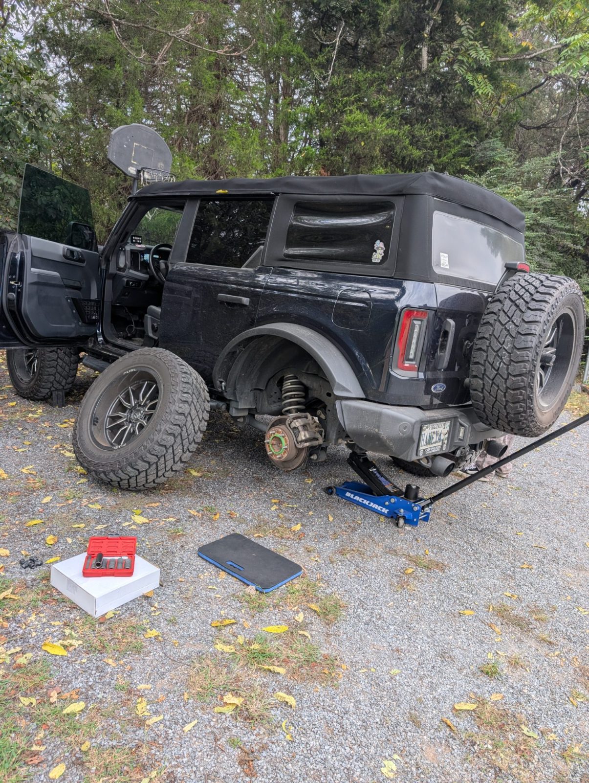 Black SUV on a floor jack in a gravel driveway with the rear wheel removed for brake work