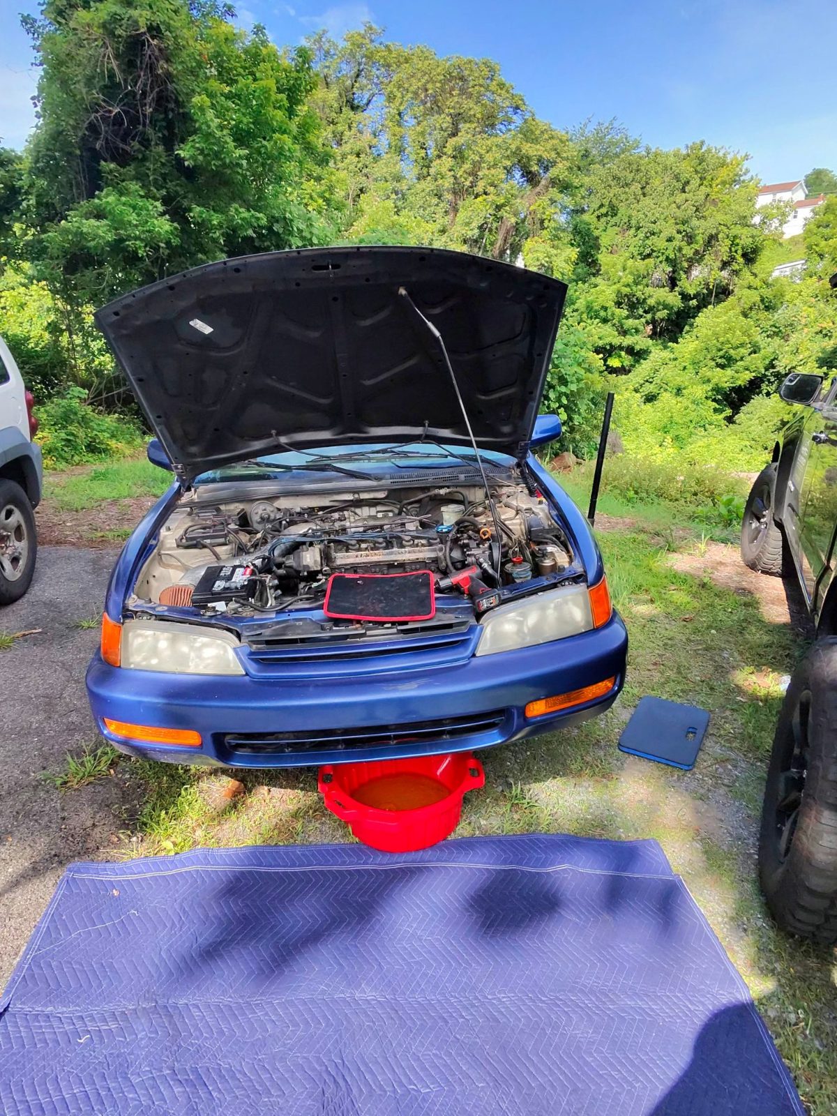 Blue car with hood up in a driveway and a red drain pan under the engine