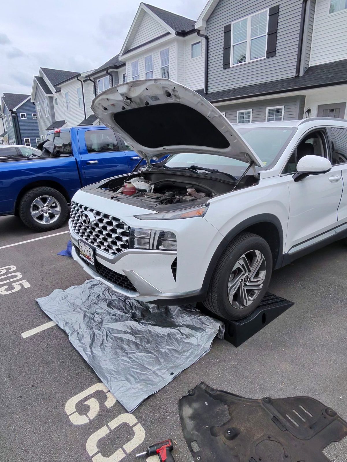 White SUV with hood up, front wheel on a ramp and splash shield removed in a parking lot